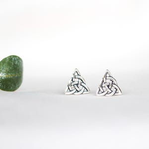 May include: A pair of silver-colored stud earrings. Each earring features a triangular Celtic knot design. The earrings are displayed on a white surface, with a green leaf in the background.