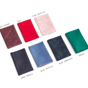 May include: A collection of small, rectangular leather goods in various colors, including red, green, and dark purple. The items are labeled with text such as "Red Lizard" and "Blue Embossed". The items are arranged on a white surface.