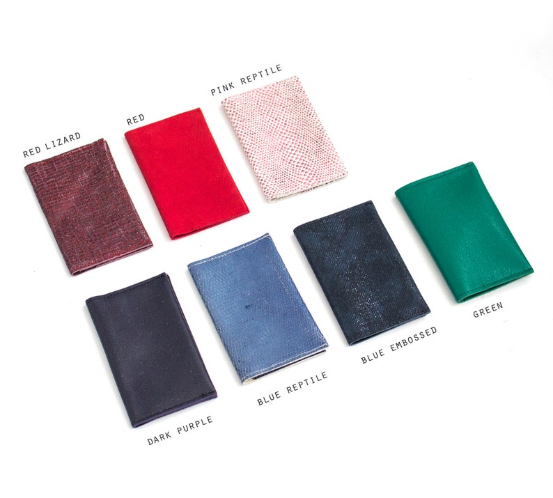 May include: A collection of small, rectangular leather goods in various colors, including red, green, and dark purple. The items are labeled with text such as "Red Lizard" and "Blue Embossed". The items are arranged on a white surface.