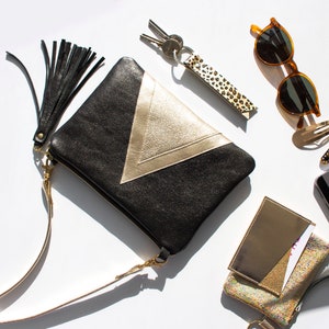 May include: Black and gold clutch with a tassel, a leopard print keychain, sunglasses, hair clips, a phone, and a small wallet. The clutch has a gold chevron design and a gold zipper. The items are arranged on a white surface.