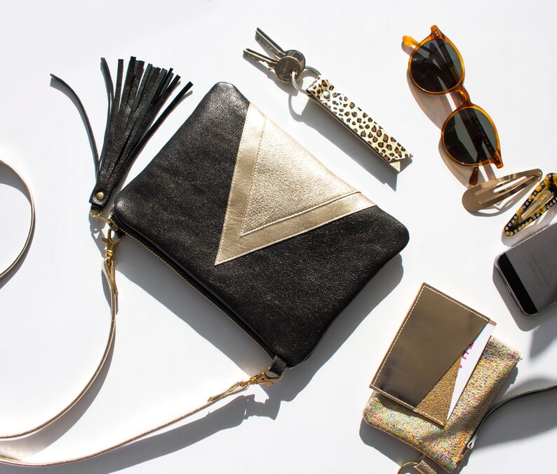 May include: Black and gold clutch with a tassel, a leopard print keychain, sunglasses, hair clips, a phone, and a small wallet. The clutch has a gold chevron design and a gold zipper. The items are arranged on a white surface.