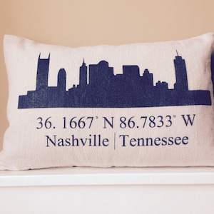 May include: A rectangular pillow with a dark blue silhouette of a city skyline. Below the skyline, the text reads "36.1667° N 86.7833° W Nashville | Tennessee". The pillow is off-white.