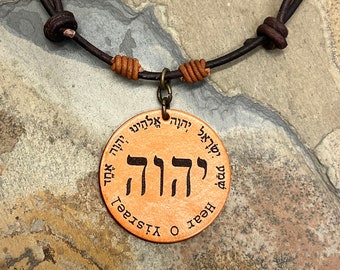 May include: A brown leather cord necklace with a round pendant. The pendant is made of wood and has the Hebrew word "L'chaim" engraved on it. The pendant also has the words "Heart" and "Visage" engraved on it.