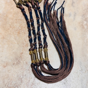 May include: Four handmade macrame keychains with bronze-colored clasps. Each keychain features a dark blue and brown twisted cord design, accented with gold-colored metal beads. The keychains are arranged on a light-colored surface.