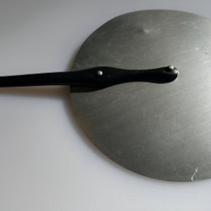 Puede incluir: Una tapa de metal plateado con un mango de plástico negro. La tapa es redonda y tiene un pequeño pomo en la parte superior.