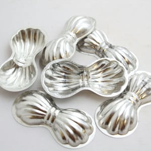 May include: A collection of six silver-colored, bow-shaped molds. Each mold has a detailed, ribbed design, with a rounded shape at each end and a narrower center. The molds are arranged on a white surface.