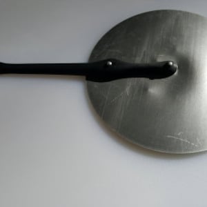 Puede incluir: Una tapa de metal plateado con un mango negro. La tapa es redonda y tiene una forma ligeramente abovedada. El mango está unido a la tapa con dos remaches.