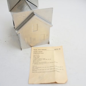 May include: A silver metal house-shaped baking pan with a recipe card for "Kasten - Festive gingerbread house". The recipe card includes ingredients and instructions.