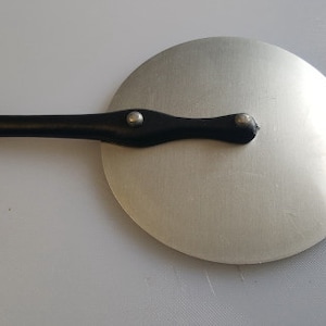 May include: A silver round lid with a black handle. The handle has a loop on one end and two small silver rivets.