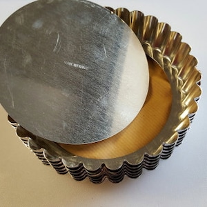 Puede incluir: Un conjunto de ocho moldes para tartas de plata acanalados con fondo extraíble. Los moldes están apilados con los fondos hacia arriba. El molde superior tiene un fondo extraíble que está colocado encima de la pila.