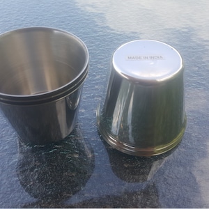 May include: Two stainless steel cups, one stacked on top of the other. The bottom cup has the words "Made in India" printed on the bottom.