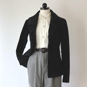 May include: A black blazer with a faux fur collar is paired with a white blouse and houndstooth pants. The outfit is accessorized with a brown belt. The ensemble is displayed on a mannequin, showcasing a classic, vintage style.