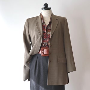 Puede incluir: Un maniquí exhibe un conjunto completo. Incluye una blazer marrón, una camisa estampada con botones, una falda lápiz gris oscuro y un cinturón de cuero marrón. El conjunto es de estilo clásico y profesional.