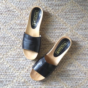 May include: A pair of Anne Klein slide sandals featuring black crocodile-embossed leather straps and light-colored wooden soles. The black footbeds display the Anne Klein logo and the text Made in Italy in gold lettering on the heel area.
