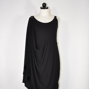 Peut inclure: Robe noire drapée sur un mannequin. La robe présente une conception à une épaule avec une silhouette asymétrique et fluide. Le tissu semble doux et comporte un détail de jupe superposée. La robe est de couleur noire unie.