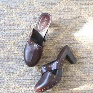 Pode incluir: Dois tamancos de couro castanho com saltos de madeira e detalhes de tachas douradas. Um tamanco tem um design de bico fechado, enquanto o outro tem um design de bico aberto. A palavra "LAUREN" é visível na palmilha de um tamanco. Os tamancos estão em um tapete tecido.