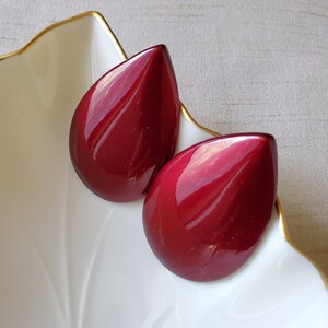 Op de afbeelding: Twee glanzende, druppelvormige oorbellen in een diepe bordeauxrode kleur. De oorbellen liggen op een witte, bladvormige schaal met een gouden rand. De oorbellen hebben een glad, reflecterend oppervlak.
