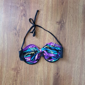 May include: A black bikini top with a halter neck tie. The cups feature a vibrant pattern of purple, teal, and gold stripes on a black background. The top is designed with a bow detail in the center.