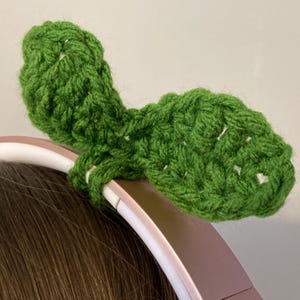 May include: Green crocheted leaf-shaped headband for headphones. The headband is made of soft yarn and is designed to fit over the top of headphones.