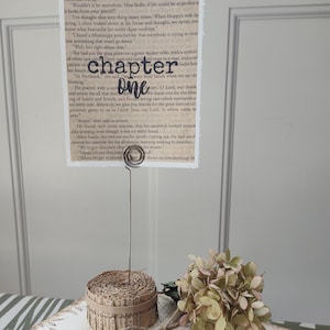 May include: A decorative display featuring a vintage book stack, a paper roll, and a tag that says "...our story." A card with the words "chapter one" is held by a wire stand. The items are in neutral tones.