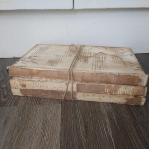 May include: A stack of four vintage-style books with brown covers and white pages. The books are tied together with twine and have a distressed, aged look.