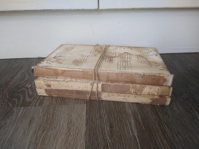 May include: A stack of four vintage-style books with brown covers and white pages. The books are tied together with twine and have a distressed, aged look.