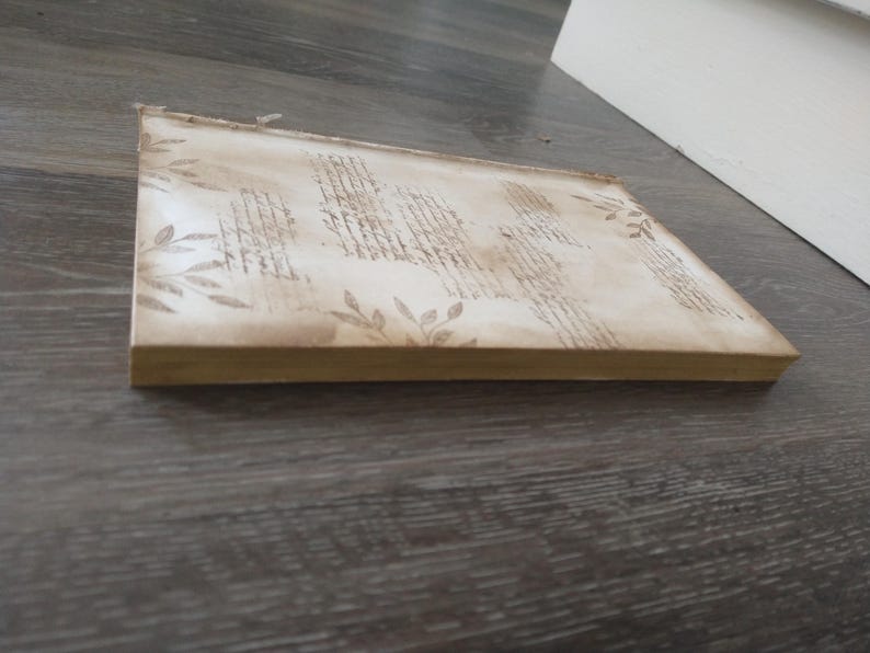 May include: A blank, off-white journal with a decorative, embossed design of leaves and lines. The journal has a light brown cover and a thick spine.