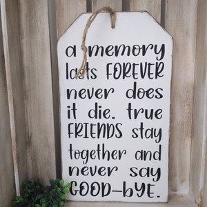 May include: White wooden tag sign with the text "a memory lasts FOREVER never does it die. true FRIENDS stay together and never say GOOD-BYE." The sign has a jute rope for hanging and a small green plant in the corner.