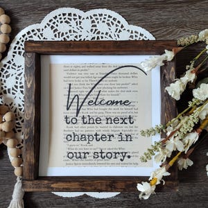 Op de afbeelding: Een vierkante donkerbruine houten lijst met een vintage boekpagina met de zwarte tekst Welcome to the next chapter in our story. De lijst ligt op een wit kanten kleedje met houten kralen en witte bloemen in de buurt.
