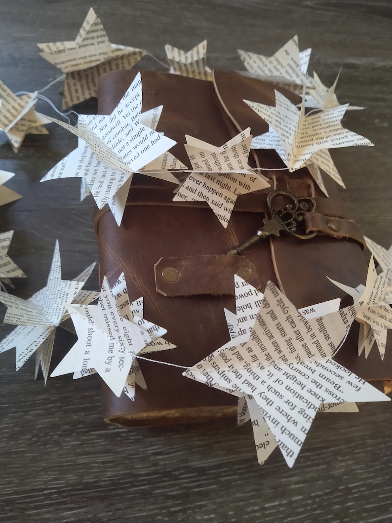 May include: A brown leather journal with a key and a string of paper stars hanging from it. The stars are made from pages of a book and have text printed on them.