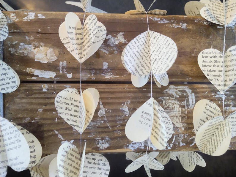 May include: A string of paper hearts made from book pages. The hearts are hanging from a rustic wooden surface. The hearts are white with black text.