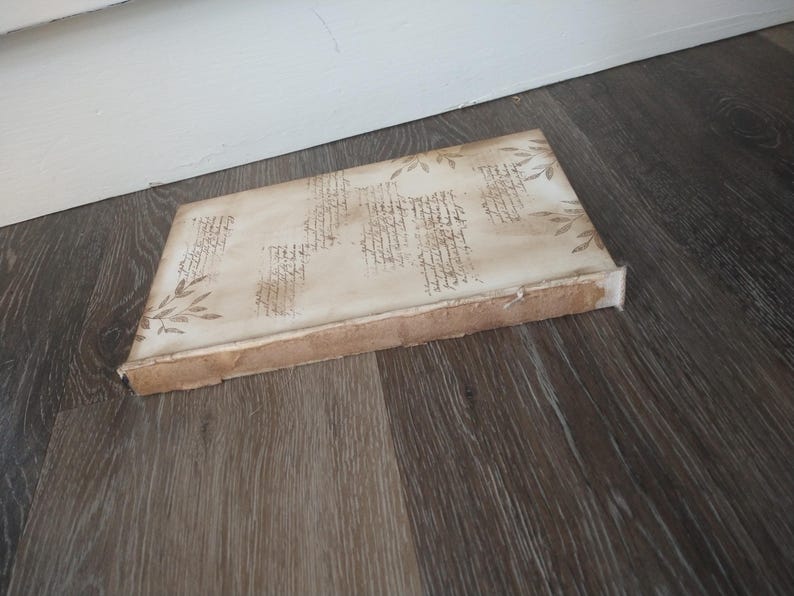 May include: A white canvas with a brown border, featuring a decorative design of handwritten script and floral elements. The canvas is lying on a wood floor.