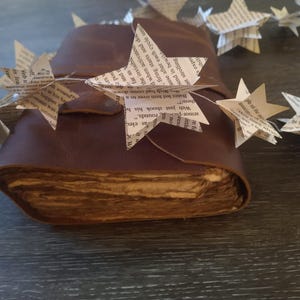 May include: A brown leather-bound journal with pages made of old, aged paper. A string of paper stars, each with text printed on them, is draped over the journal.
