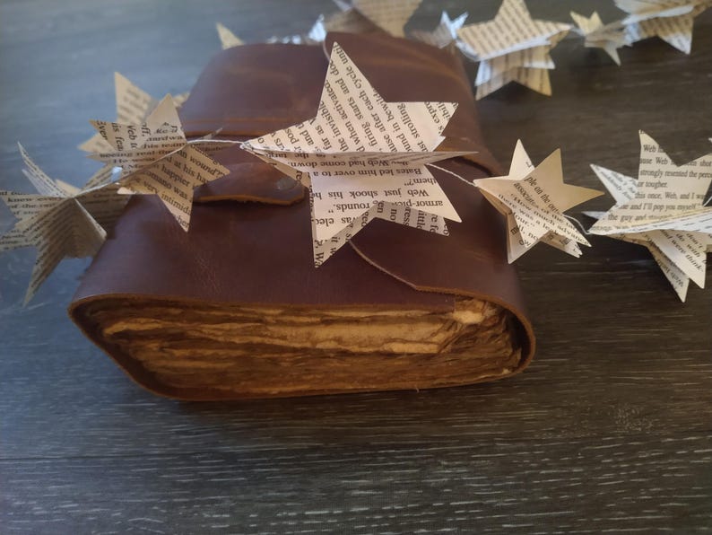 May include: A brown leather-bound journal with pages made of old, aged paper. A string of paper stars, each with text printed on them, is draped over the journal.