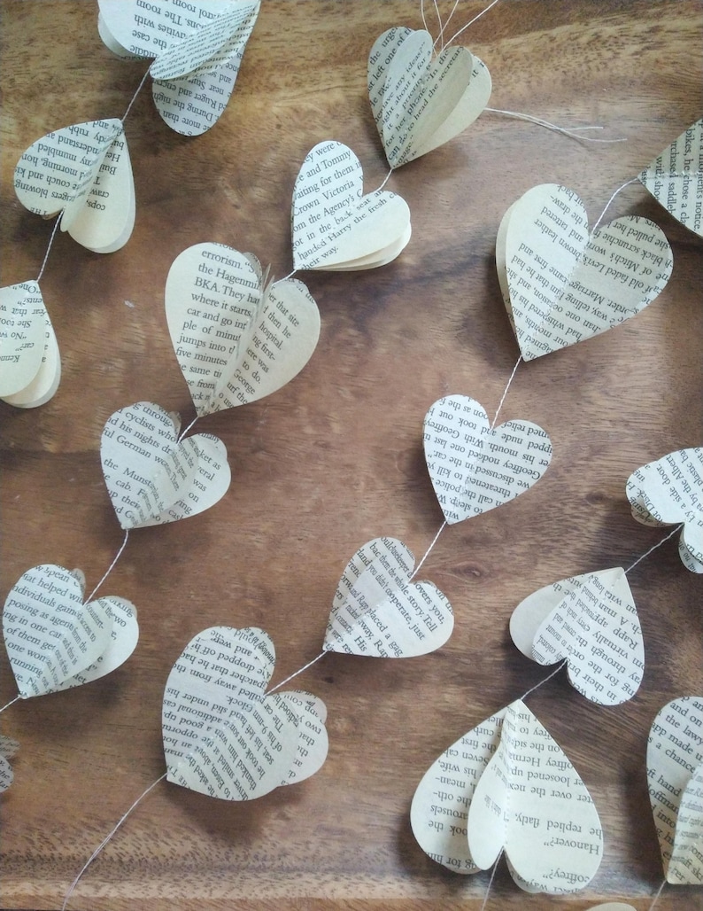May include: A garland of paper hearts made from recycled book pages. The hearts are strung together with white thread and hang on a wooden surface.