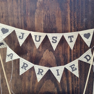 Puede incluir: Adorno para tarta con la frase "Just Married" en banderines triangulares hechos con páginas de libro. Los banderines tienen corazones negros y letras negras. Palillos de madera sostienen la pancarta.