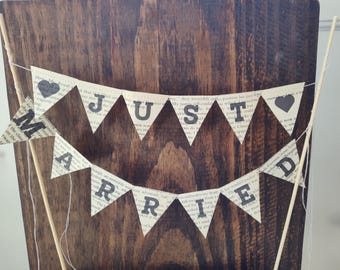 May include: A two-tiered banner cake topper with the words "Just Married" printed on triangular flags. The flags are made from book pages and accented with black hearts. Wooden dowels support the banner.