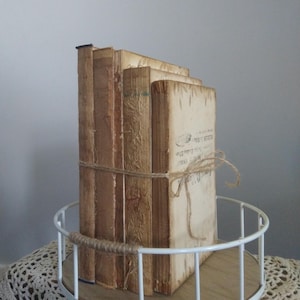 Puede incluir: Una pila de cuatro libros antiguos con cubiertas marrones y crema, atados con cuerda, sobre una bandeja de metal blanco con base de madera.