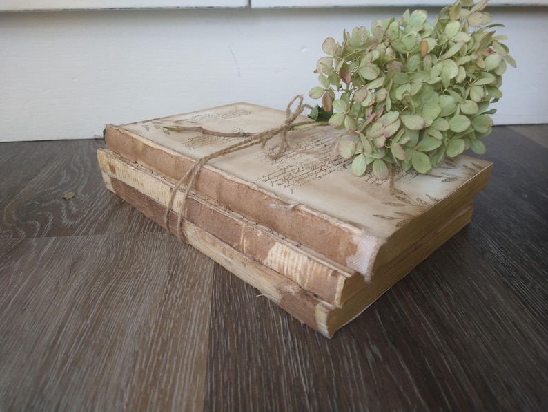 May include: A stack of three vintage books with a rustic brown cover and white pages. The books are tied together with twine and decorated with dried flowers.