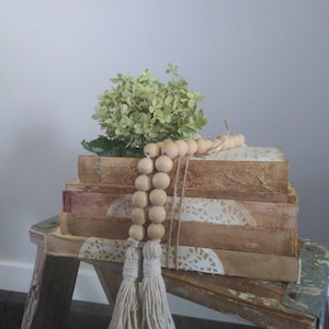 May include: A stack of vintage books with lace trim and a wooden bead garland, decorated with a sprig of dried hydrangeas. The books are arranged on a rustic wooden stool.