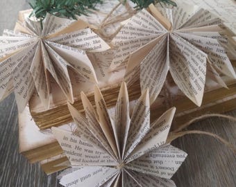 May include: A garland of book page ornaments, each folded into a star shape, hangs from a twine string. The ornaments are made from old book pages and are strung together with twine. The garland is hanging on a stack of old books.