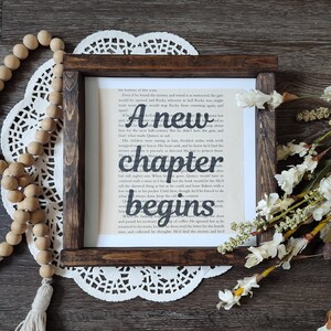 May include: A square wooden frame with a dark brown finish containing a page from a book with the black text A new chapter begins printed over the centre. The frame is surrounded by a white lace doily, wooden beads, and white flowers.