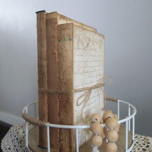 Könnte beinhalten: Drei Bücher im Vintage-Stil, die auf einem weißen Metalltablett mit Holzbasis gestapelt sind. Die Bücher sind in braunes Papier gebunden, das einen abgenutzten Look hat, und haben handschriftlichen Text auf den Einbänden. Eine Schnur aus Holzperlen mit einem weißen Quasten hängt am Tablett.