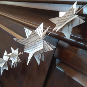 May include: A garland of paper stars made from book pages hangs on a dark wood surface. The stars are strung together with white thread and have text printed on them.