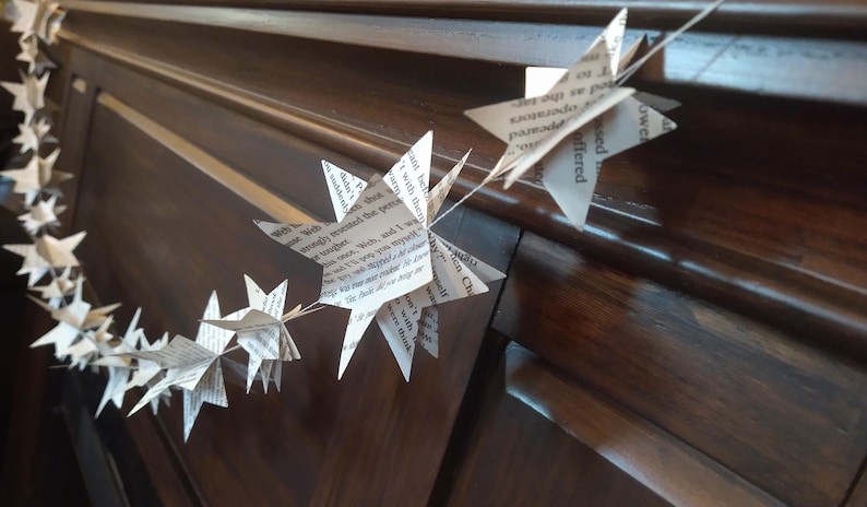 May include: A garland of paper stars made from book pages hangs on a dark wood surface. The stars are strung together with white thread and have text printed on them.