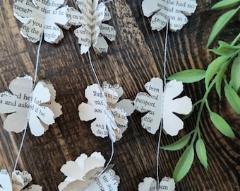 May include: A garland of paper flowers made from book pages. The flowers are strung together with white thread and hang against a wooden background. The flowers are white with black text from the book pages.