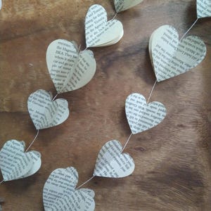 May include: A string of paper hearts made from book pages, hanging on a wooden surface. The hearts are connected by a thin string and are decorated with printed text.