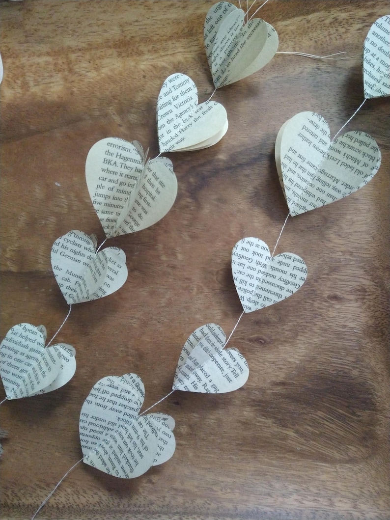 May include: A string of paper hearts made from book pages, hanging on a wooden surface. The hearts are connected by a thin string and are decorated with printed text.
