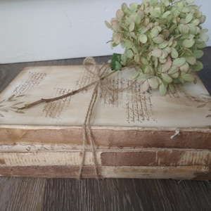 May include: A stack of three vintage books with brown covers and white pages. The books are tied together with twine and decorated with dried flowers.