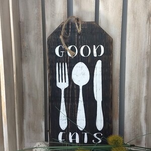 Puede incluir: Un letrero de madera rústico con las palabras "GOOD EATS" pintadas en blanco. El letrero también presenta una silueta blanca de un tenedor, una cuchara y un cuchillo.
